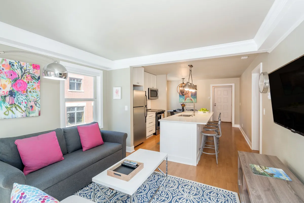A view of an open-plan living area showing a comfortable sofa with pink cushions, a well-appointed kitchen island, and colorful artwork. The kitchen is integrated with the living space, emphasizing a casual, social atmosphere where cooking and connecting blend seamlessly.