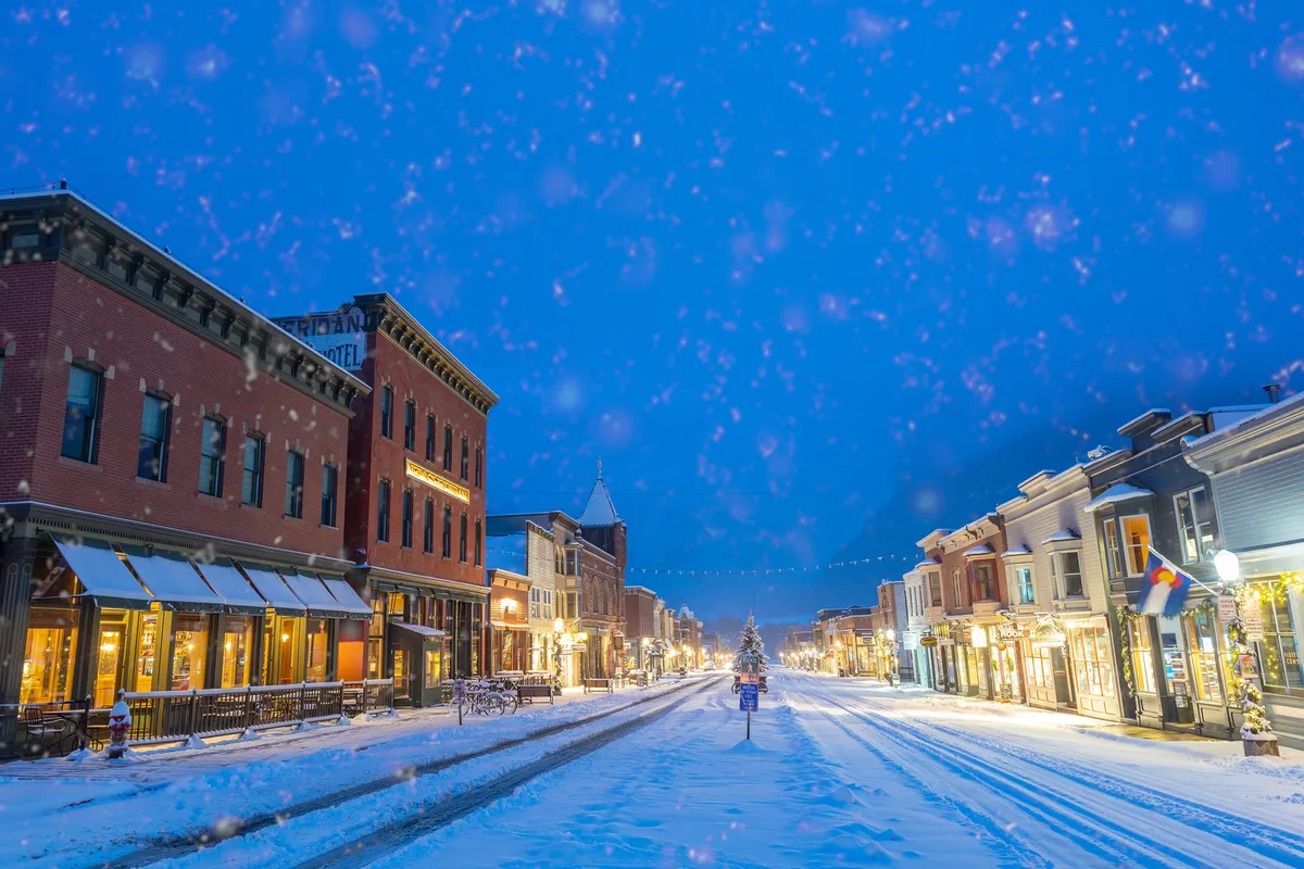 A picturesque snow-covered street lined with decorated storefronts and a glowing Christmas tree in the background. People stroll along the sidewalk, immersed in a festive, cozy atmosphere that highlights the town's charm during events and celebrations.