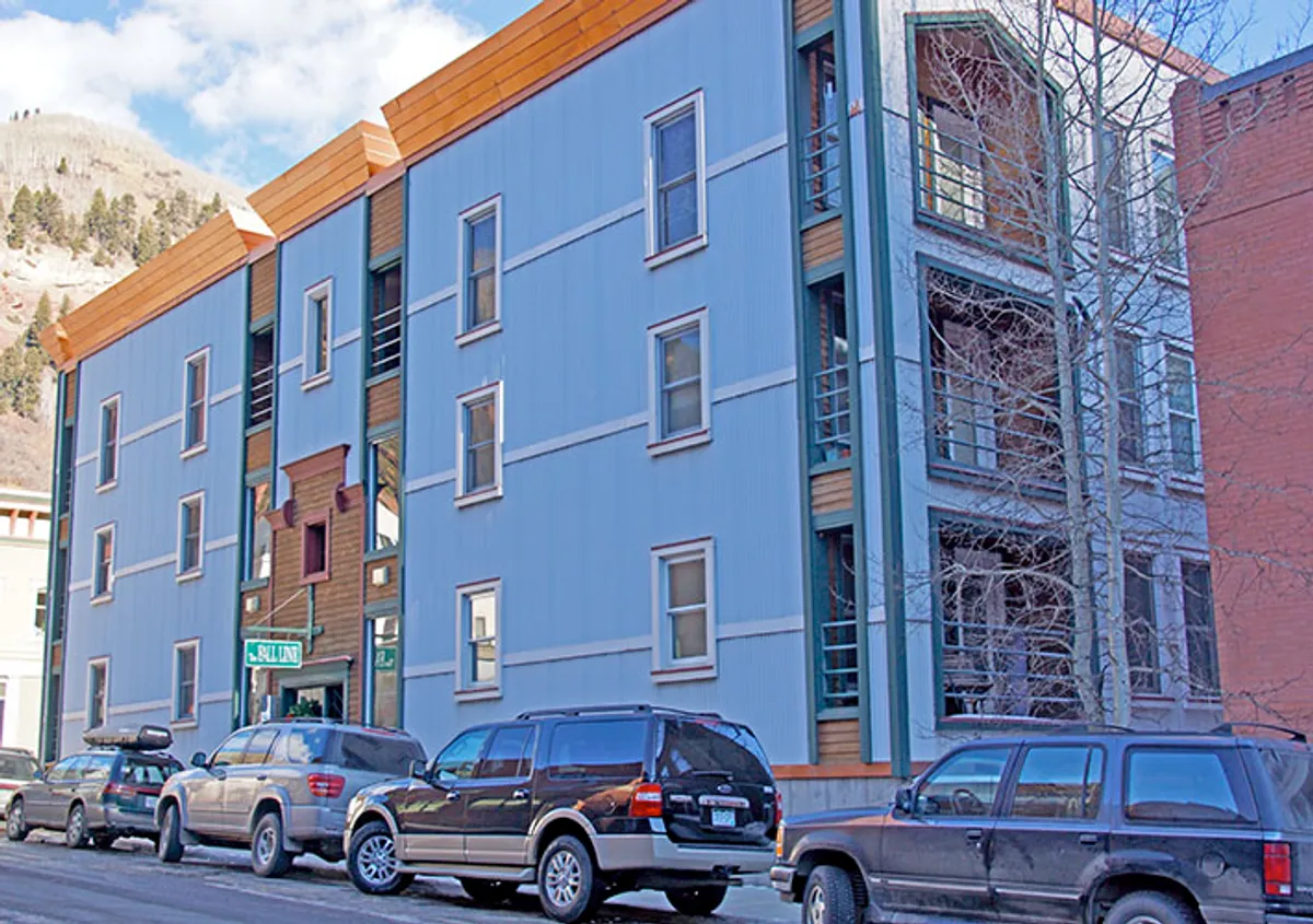 The image features the exterior of the building with a unique architectural design and vibrant colors, framed by parked vehicles and a stunning mountain backdrop. The lively urban atmosphere suggests proximity to local attractions and the energy of Telluride's Main Street.