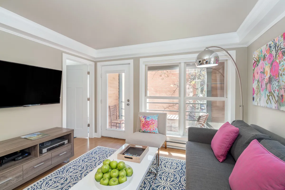 This image features a comfortable living area with a sofa, flat screen television, vibrant artwork, green apples on a table, and a balcony door letting in natural light. The room feels welcoming and bright, inviting both socializing and relaxation. The visible balcony door hints at access to outdoor space and mountain views.