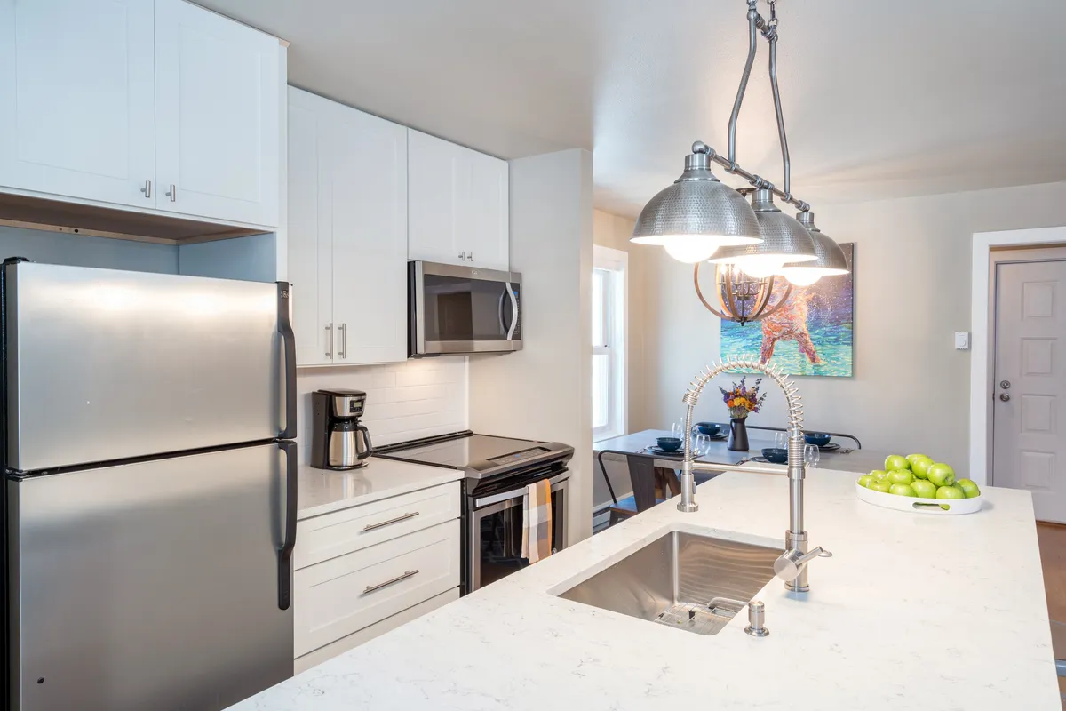 A spacious, welcoming kitchen featuring a modern kitchen island, gleaming stainless steel appliances, a bowl of fresh fruit, and tasteful artwork on the wall. The space is clearly set up for both cooking and entertaining, with clean countertops and plenty of room to gather.