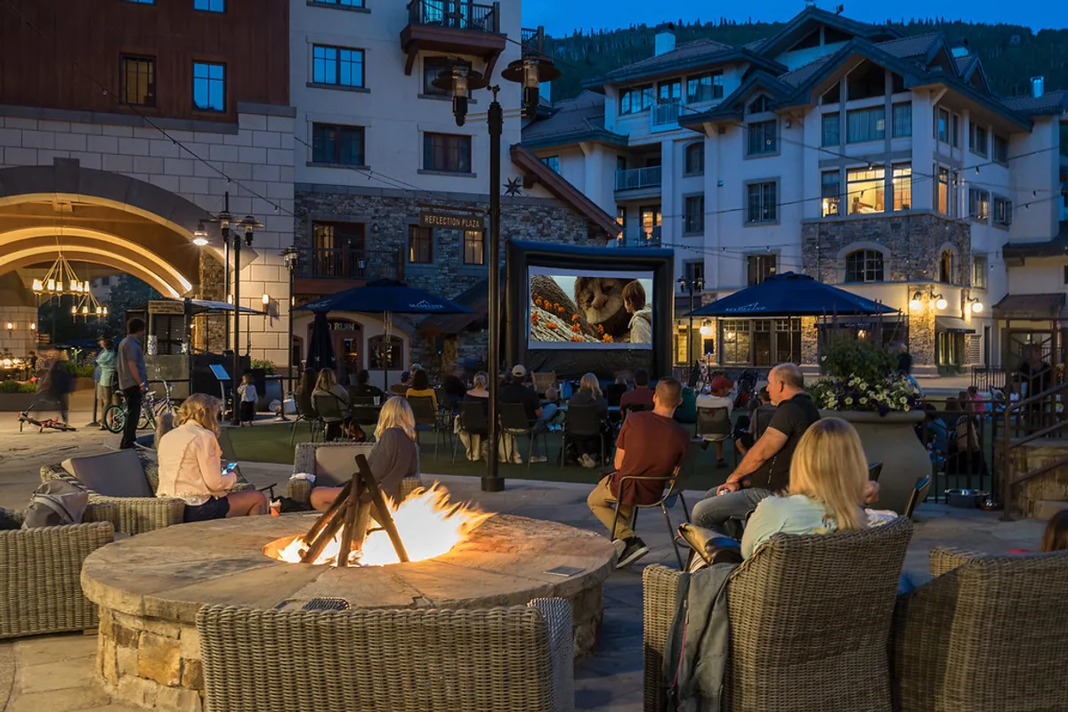 A lively outdoor area featuring a fire pit, an outdoor movie screen set up for entertainment, cozy seating, and the building facade in the background. People are seen relaxing and socializing, enjoying a communal atmosphere under the open sky.