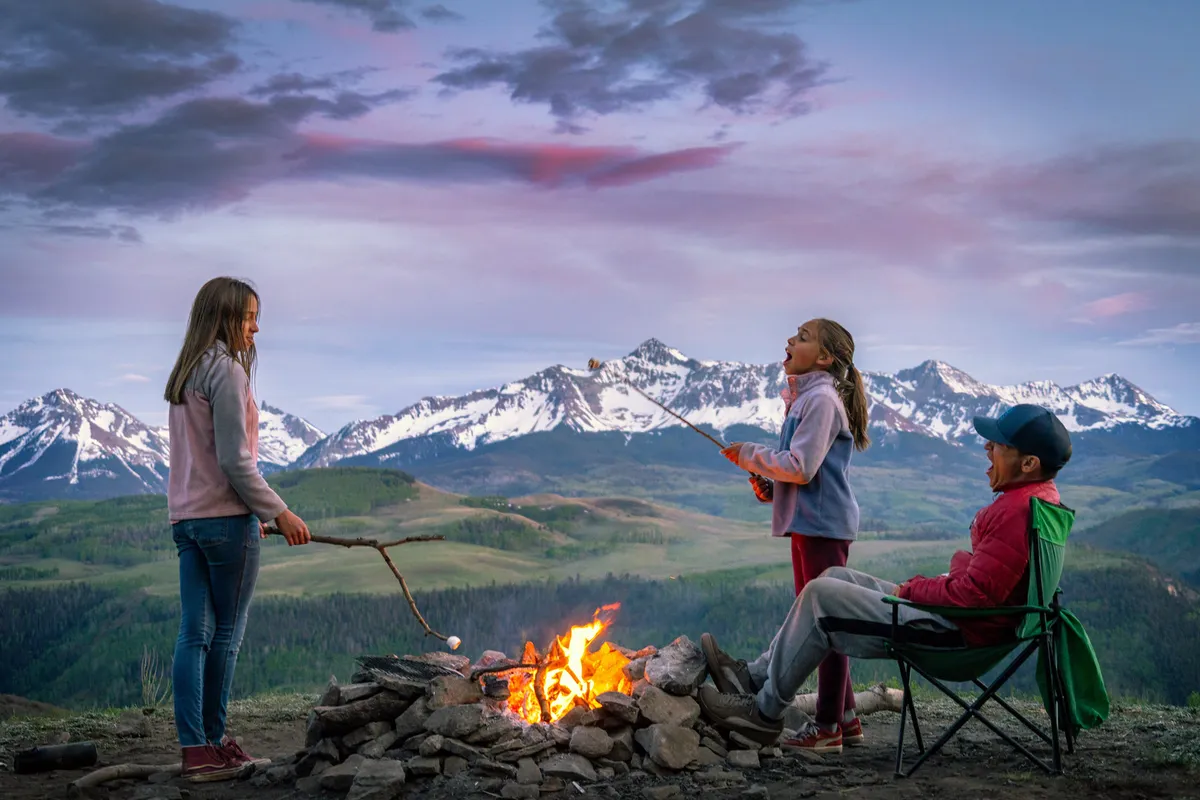 Children roast marshmallows over a campfire at the base of a mountain range while a man relaxes nearby, creating a warm, joyful family moment surrounded by nature.