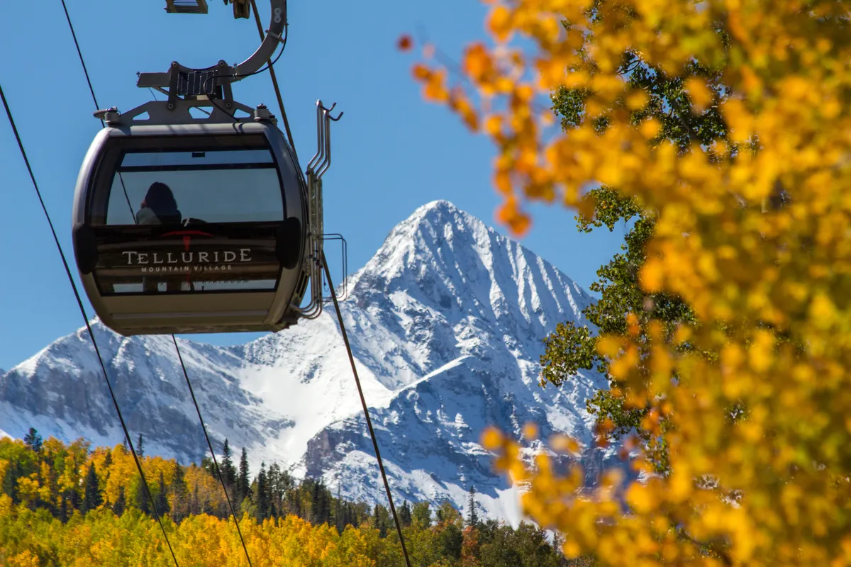 A breathtaking exterior scene featuring the gondola unmistakably connecting to mountain adventure, with snow-capped peaks in the background and vibrant autumn foliage in the foreground. The image vividly showcases the region's dual seasonal appeal for both skiers and hikers, and the gondola signals direct access to mountain exploration.