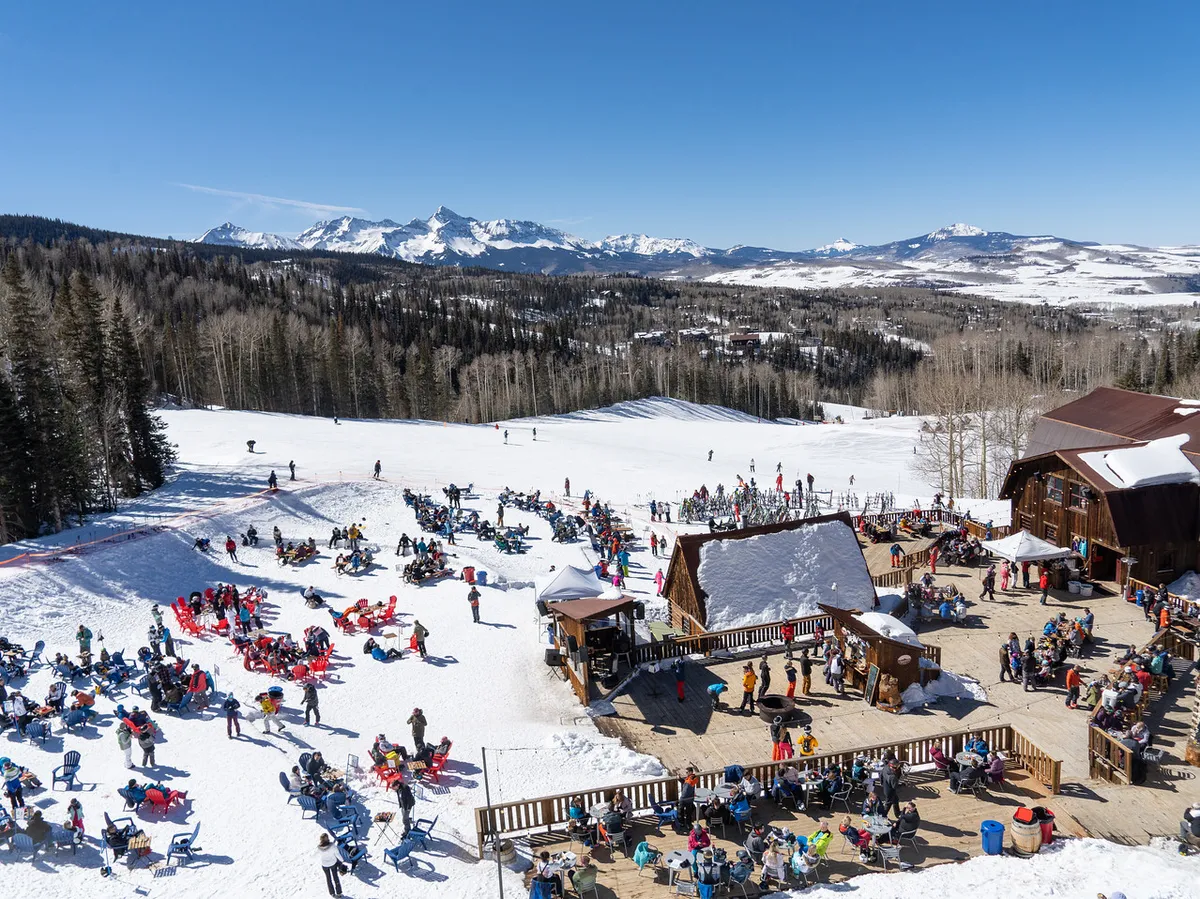 The image shows skiers on the slopes, a lively crowd enjoying an outdoor seating area, and snow-covered mountains forming a dramatic backdrop. Wooden structures and a ski lodge suggest a welcoming spot for après-ski gatherings or meals with friends. The energy of people socializing and the beautiful alpine scenery establish a vibrant, communal atmosphere.