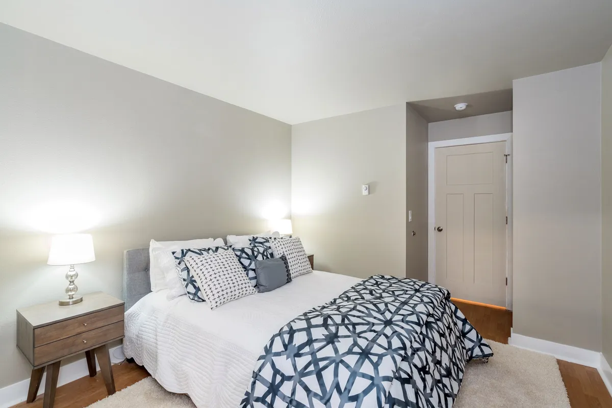 A cozy bedroom with a bed dressed in patterned bedding and a nightstand with a lamp, creating a warm, intimate space. The setting is perfect for relaxing, reading, or winding down for the night.