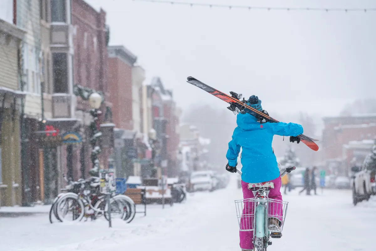 A person rides a bicycle down a snow-covered street, surrounded by ski equipment and buildings. The scene captures the spirit of exploring town in winter, with hints of both activity and relaxation. The urban setting and visible buildings evoke easy access to local bars, restaurants, and community events.