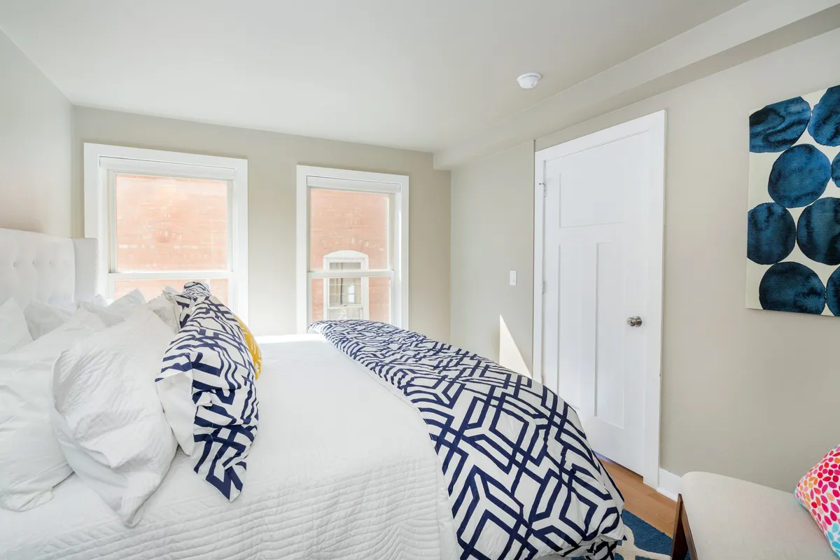 A serene bedroom featuring a bed with patterned bedding, large windows letting in natural light, and tasteful wall art. The atmosphere is peaceful and inviting, suggesting a restful night’s sleep after a long day.