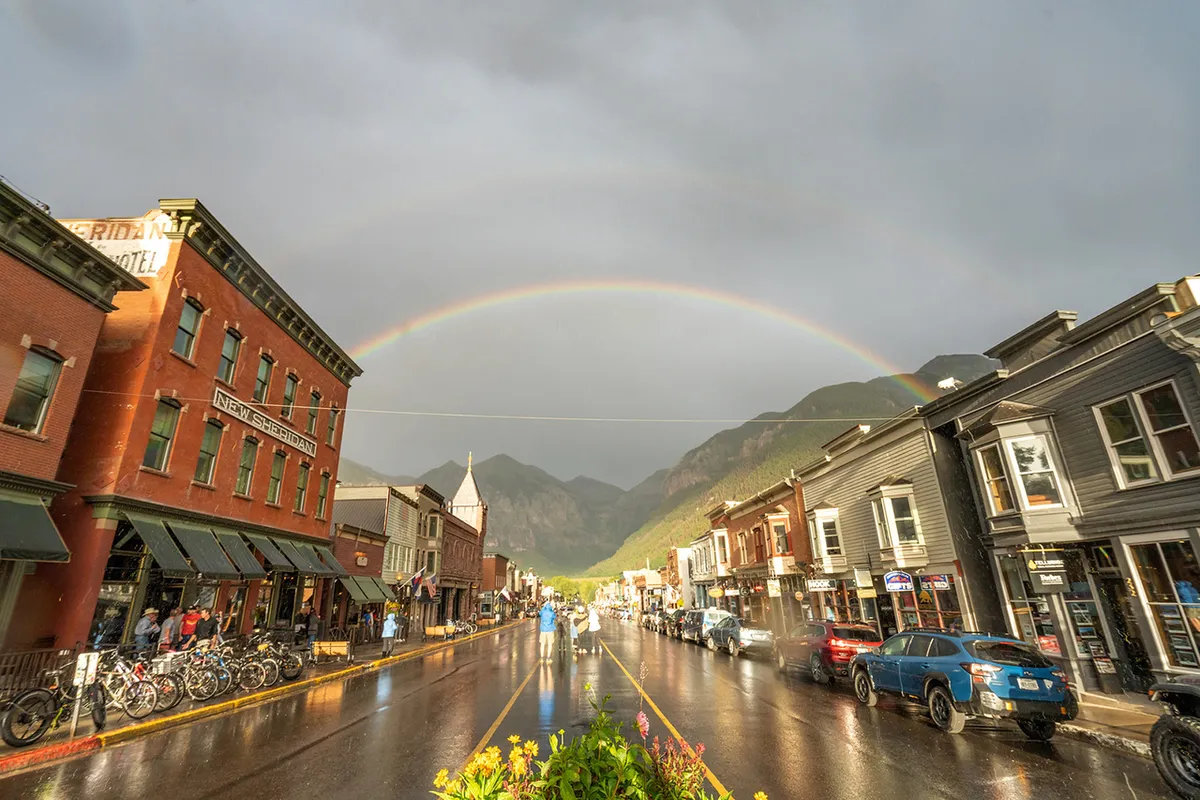A charming Telluride street scene showcasing historic buildings, a rainbow arching overhead, wet pavement that hints at recent rain, and a mountainous backdrop. The composition invites exploration and a sense of community.